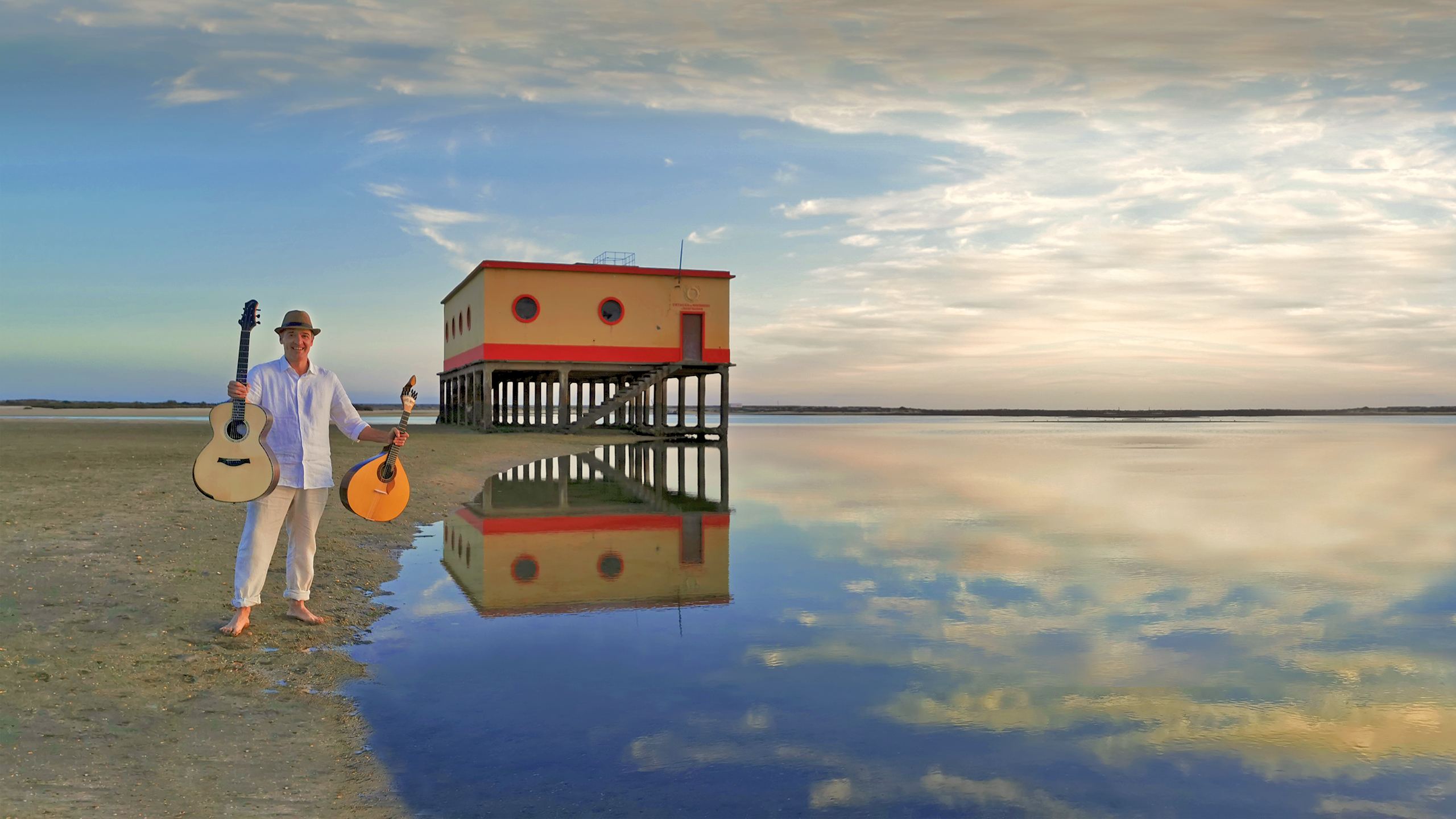 João Cuña barefoot on the Ria Formosa sandflat, holding an acoustic guitar in one hand and a Portuguese guitar in the other, in front of the lifeboat station mirrored in the water at dawn